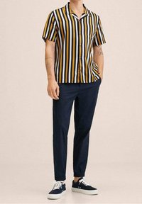 Camisa de manga corta con botones presenta rayas verticales en amarillo mostaza, negro y blanco. Combinada con pantalones azul marino y zapatillas oscuras.