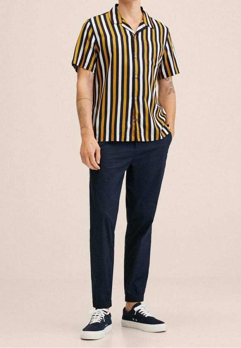 Camicia a maniche corte con bottoni presenta strisce verticali in giallo senape, nero e bianco. Abbinata a pantaloni blu navy e sneaker scure.