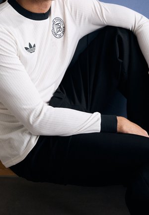 Mann trägt weißes Langarmhemd mit schwarzem Kragen und Deutschem Fußball-Bund-Logo, sitzt mit einem angewinkelten Knie, trägt schwarze Hose.