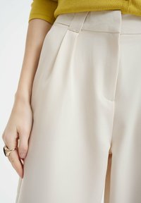 Beige wijde broek met plooien bij de taille en zijzakken. Glad, lichtgewicht stof met een subtiele glans. Hand met een zichtbare ring.