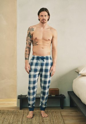 Mann mit Tattoos steht barfuß in blau-weiß karierter Schlafanzughose in einem Schlafzimmer mit beigen und grauen Wänden.