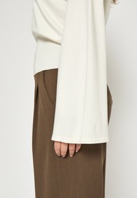 Krémový top s dlhým rukávom a voľným strihom, so širokým lemom, v kombinácii s hnedými plisovanými nohavicami, ktorý predvádza hladkú textúru látky.