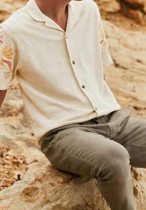 Persoon in een beige kortemouwen overhemd met bloemenmouwen en grijze broek, zittend op een rotsachtige ondergrond in de open lucht.