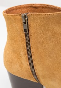 Botín de ante en color topo con un tacón de madera oscura; cuenta con una cremallera lateral con tirador metálico, mostrando una textura suave y líneas limpias.