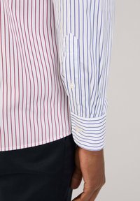 Camicia a righe con strisce verticali alternate rosse e blu su sfondo bianco, con polsino arrotondato e dettaglio dei bottoni.