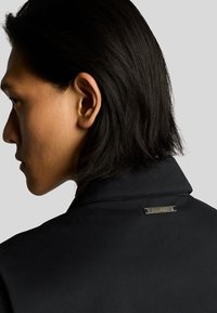 Chaqueta negra con un cuello estructurado, que presenta una etiqueta metálica en la parte posterior del cuello y una textura lisa, mostrada desde un ángulo lateral.