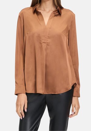 Vrouw draagt een bruine blouse met lange mouwen en V-hals en zwarte broek, staand tegen een effen witte achtergrond.