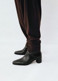 Zwarte leren enkellaarsjes met een vierkante neus en een blokhak, gecombineerd met losse bruine broeken. Gladde textuur en minimalistisch ontwerp.