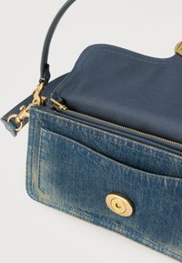 Μικρό μπλε τζιν τσάντα με χρυσό κούμπωμα, φερμουάρ και μπλε δερμάτινο καπάκι, μισάνοιχτη που δείχνει εσωτερικά διαμερίσματα και σημείο στερέωσης λουριού.