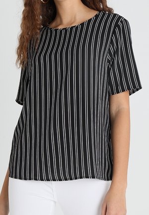 Woman wearing black short-sleeve top with vertical white stripes and white pants, standing against a plain light background.