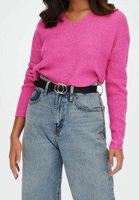 Fuchsiafarbenes, geripptes Sweatshirt mit V-Ausschnitt, kombiniert mit hellblauen, hoch taillierten Jeans. Dazu ein schwarzer Gürtel mit runder silberner Schnalle.