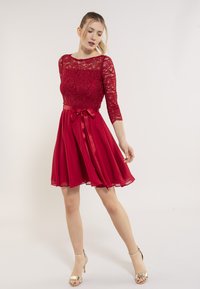 Robe rouge avec un corsage en dentelle, des manches trois-quarts et une jupe en chiffon fluide. Présente une taille en satin avec un détail de nœud.