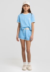 Niebieska bawełniana koszulka z białym wykończeniem i logo, zestawiona z niebieskimi szortami z elastycznym pasem. Całość uzupełniają białe trampki i czapka.