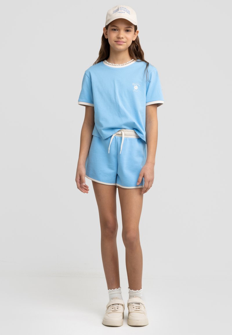 Niebieska bawełniana koszulka z białym wykończeniem i logo, zestawiona z niebieskimi szortami z elastycznym pasem. Całość uzupełniają białe trampki i czapka.