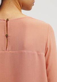 Brzoskwiniowa bluzka o miękkiej teksturze, z rozcięciem z tyłu i dwoma ozdobnymi guzikami przy dekolcie. Casualowy design z luźnym krojem.