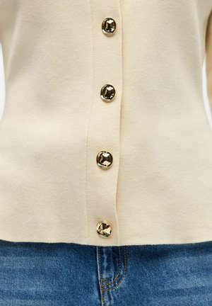 Cardigan crème à quatre boutons dorés brillants, boutonné sur un jean en denim bleu, montrant le détail de la taille.