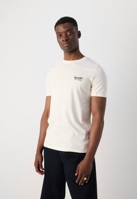 Tricou din bumbac de culoare crem, cu mâneci scurte, având un grafic verde și text, asortat cu pantaloni de culoare bleumarin.