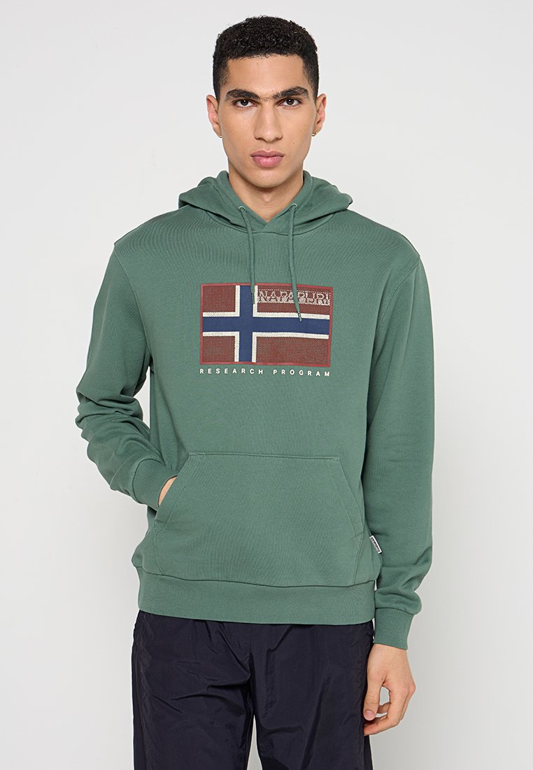 Junger Mann in einem grünen Kapuzenpullover mit einer Fronttasche und einer rot-weißen-blauen Fahnengrafik mit der Aufschrift "Napapijri Research Program".
