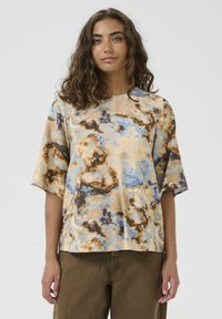 Frau mit lockigem braunem Haar, die ein lockeres, kurzärmliges Batikshirt in Beige, Blau und Braun sowie olivgrüne Hosen trägt, vor einem einfarbigen Hintergrund.