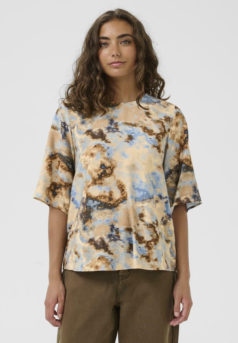 Frau mit lockigem braunem Haar, die ein lockeres, kurzärmliges Batikshirt in Beige, Blau und Braun sowie olivgrüne Hosen trägt, vor einem einfarbigen Hintergrund.