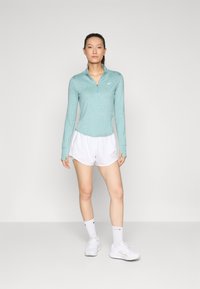Hellblaues, langärmliges Sportoberteil mit einem halben Reißverschluss, kombiniert mit weißen Shorts. Glatte Textur, athletic fit und Nike-Logo auf der Brust.