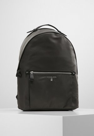 Zwarte Michael Kors rugzak met voorvak met ritssluiting en bovenhandvat, tentoongesteld op een wit voetstuk tegen een lichtgrijze achtergrond.