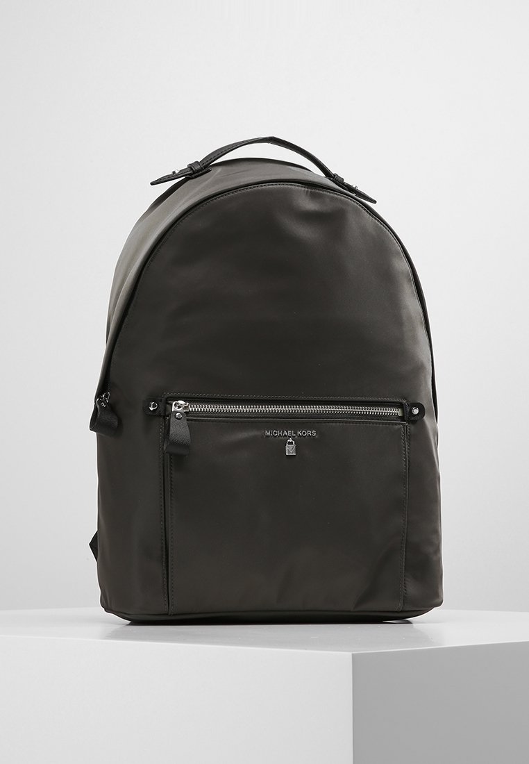 Sac à dos noir Michael Kors avec poche zippée à l'avant et poignée supérieure, exposé sur un piédestal blanc sur fond gris clair.