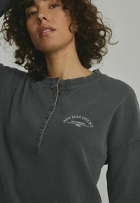 Grå sweatshirt med kvart-knap halsudskæring, blød tekstur og broderet tekst med "NEW YORK CITY NY Manhattan 1985."