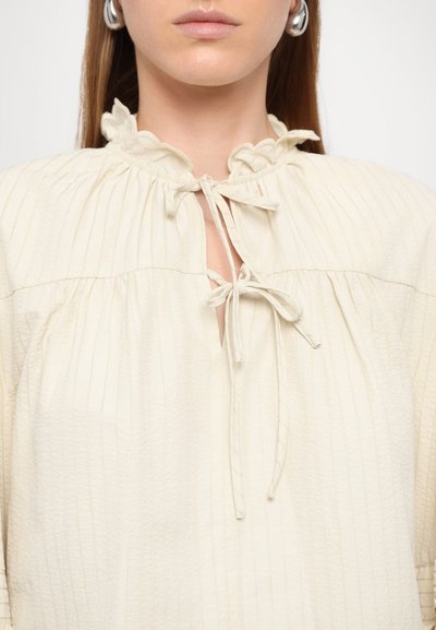Cream-colored blouse with vertical ribbed texture, ruffled collar, and tie closure at the neck. Loose-fitting design with gathered accents.