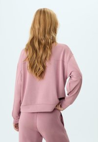 Donna con lunghi capelli mossi, che indossa un felpa rosa oversize e pantaloni coordinati, in piedi con una mano sulla vita, di spalle rispetto all'osservatore.