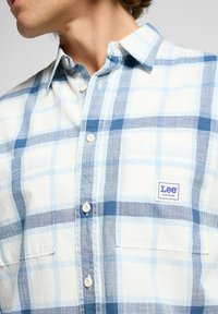 Camisa de cuadros azul claro de botones hecha de algodón. Presenta un bolsillo en el pecho y una pequeña etiqueta con el logo "Lee" en el lado izquierdo.