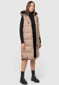 Gilet long beige rembourré avec capuche, doté d'une fermeture éclair à l'avant, de poches latérales et d'une finition texturée. Porté sur un col roulé noir et une jupe.