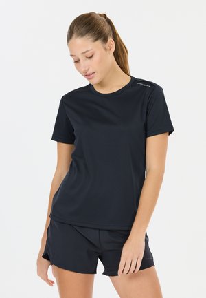 Ung kvinde med hestehale iført sort atletisk T-shirt med korte ærmer og matchende shorts, kigger nedad mod en ensfarvet baggrund.