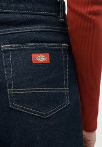Τσέπη από τζιν παντελόνι με ορατό λογότυπο Dickies, φορεμένο με μακρυμάνικη, ριμπ μπλούζα σε σκουριά χρώμα στον βραχίονα.