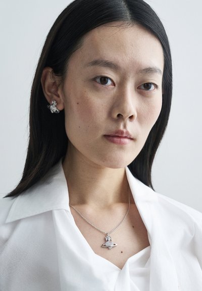 Ασημένια κοσμήματα με κρεμαστό που μοιάζει με πλανήτη και έχει λαμπερά στοιχεία, φορεμένο σε μια λεπτή αλυσίδα. Το μοντέλο φοράει ένα λευκό πουκάμισο με γιακά.