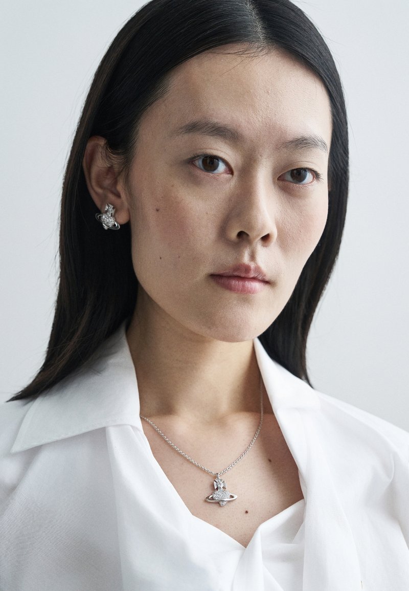 Silberner Schmuck präsentiert einen Anhänger, der einem Planeten ähnelt und mit funkelnden Akzenten versehen ist, getragen an einer zarten Kette. Das Model trägt ein weißes Hemd mit Kragen.