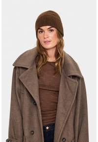 O femeie poartă o căciulă tricotată maro, o cămașă maro, o haină maro supradimensionată și blugi negri, pe un fundal deschis simplu.