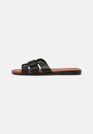 Sandalias planas negras con tiras cruzadas, hechas de material suave. Diseño minimalista con una suela interior marrón contorneada y una suela exterior negra plana.