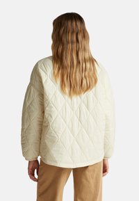 Krémová prošívaná bunda s diamantovým vzorem, volný střih a uvolněná ramena. Hladká pletenina z bavlny, v kombinaci s béžovými kalhotami.