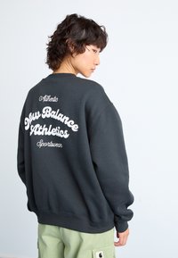 Svart hettegenser med "New Balance Athletics" og "Authentic Sportswear" trykt i hvitt på baksiden, med en avslappet passform og ribbestrikkede mansjetter.