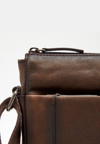 Brown leather bag featuring a textured finish, a zip closure, and metal hardware. The design includes a side pocket and a strap attachment.