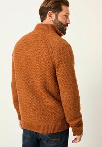 Texturierter orangefarbener Pullover mit hohem Kragen, gerippten Bündchen und Saum, der ein geometrisches Muster auf einem glatten Stoffhintergrund zeigt.