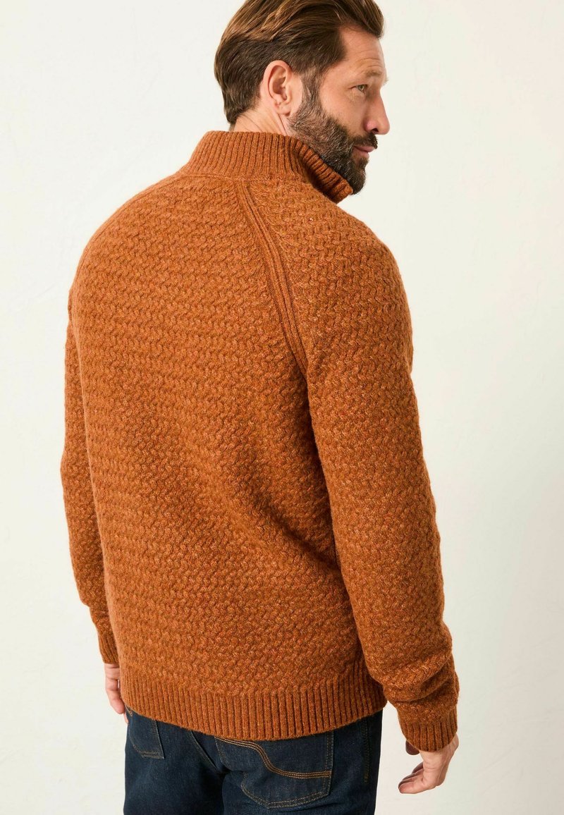 Texturierter orangefarbener Pullover mit hohem Kragen, gerippten Bündchen und Saum, der ein geometrisches Muster auf einem glatten Stoffhintergrund zeigt.