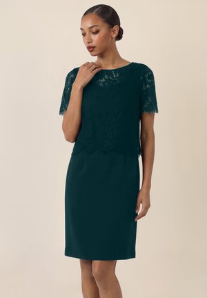 Frau mit hochgestecktem Haar, die ein dunkelgrünes knielanges Kleid mit kurzen Spitzenärmeln und Spitzenstoff auf dem Oberteil trägt, steht vor einfachem Hintergrund.