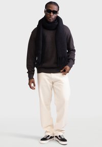 Sort strikket trøje med ribbet tekstur, parret med cremede, bred-benede bukser og sorte sneakers. Accessoriseret med et sort tørklæde og solbriller.