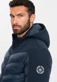 Marineblaues Pufferjacke mit Kapuze, mit einem gesteppte Design, matter Textur und einem runden Logo-Akzent am linken Ärmel. Mit Reißverschluss.