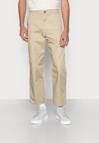 Muž v béžových kalhotách rovného střihu, bílých ponožkách a světle šedých teniskách, stojící na pozadí čistě bílé stěny.