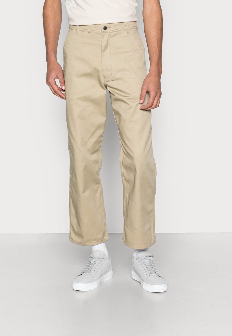 Muž v béžových kalhotách rovného střihu, bílých ponožkách a světle šedých teniskách, stojící na pozadí čistě bílé stěny.