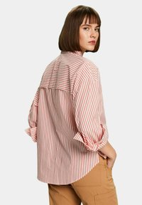 Camicia a righe rosse e bianche con una vestibilità rilassata, maniche arrotolate e dettaglio sul retro. Il tessuto sembra leggero e traspirante.
