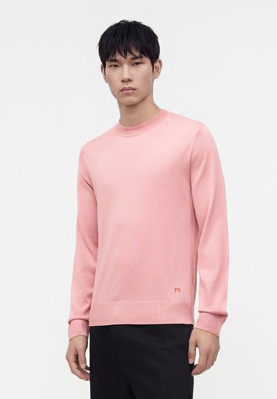 Jeune homme aux cheveux courts noirs portant un pull col rond rose clair et un pantalon noir, debout devant un fond blanc uni.